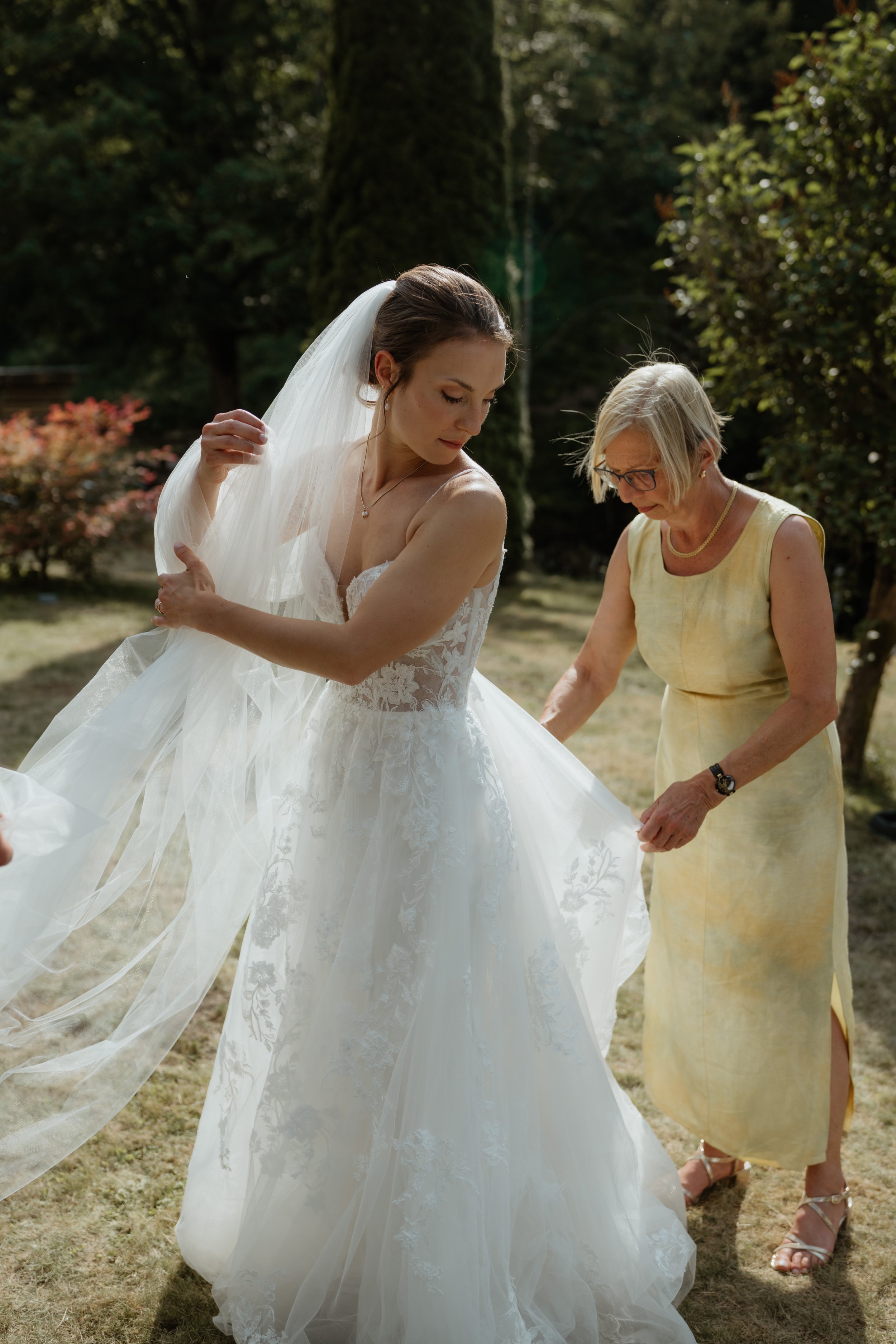 Eine junge Frau in einem weißen Hochzeitskleid, begleitet von einer älteren Frau in einem gelben Kleid, steht im Freien auf einer Wiese, umgeben von Bäumen. Die junge Frau trägt ein Vlies und hat lange braune Haare, während die ältere Frau blonde Haa