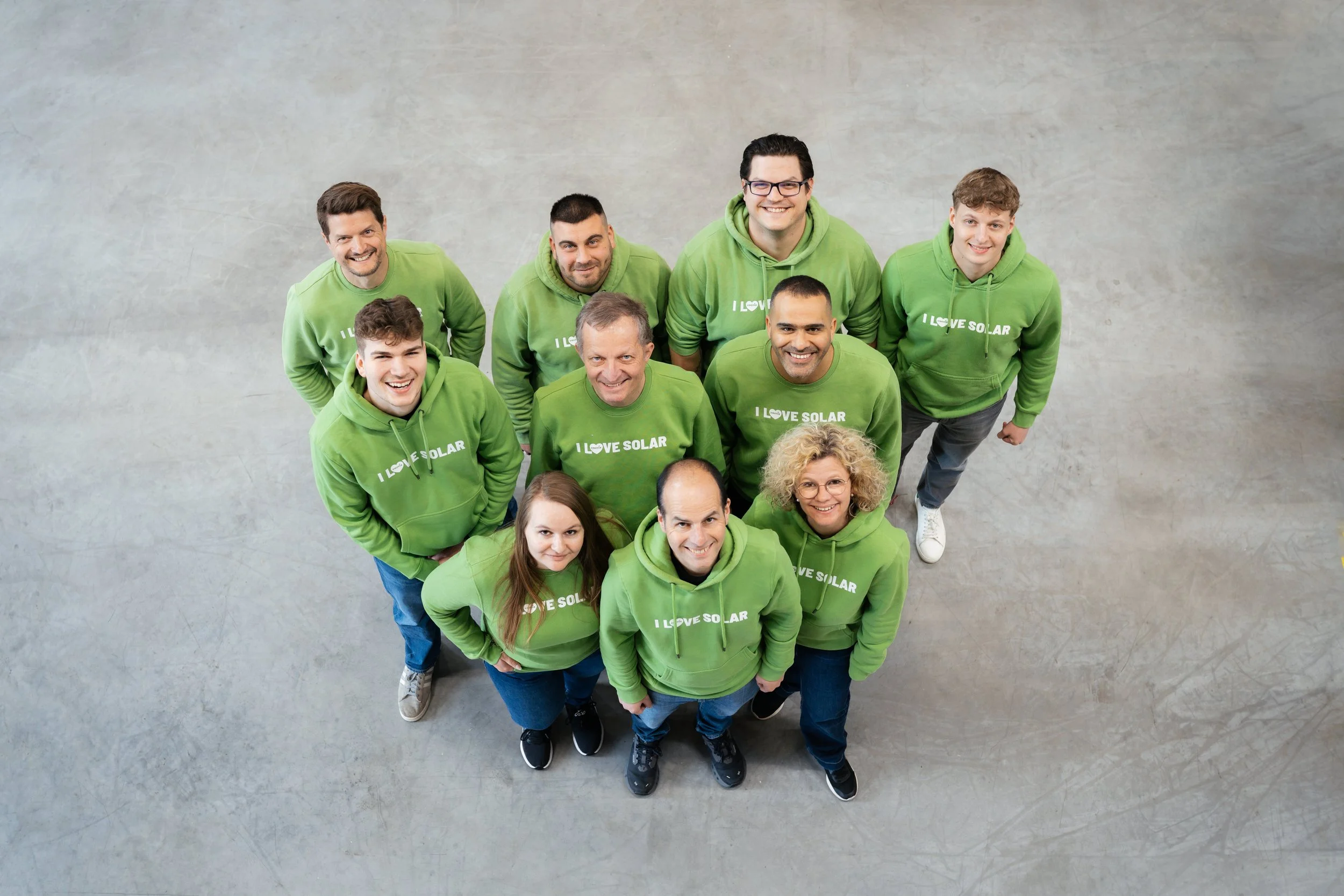 Eine Gruppe von Menschen trägt grüne Hoodies mit dem Aufdruck ‚I love solar‘, sie lächeln und schauen nach oben, vor einem neutralen grauen Hintergrund.
