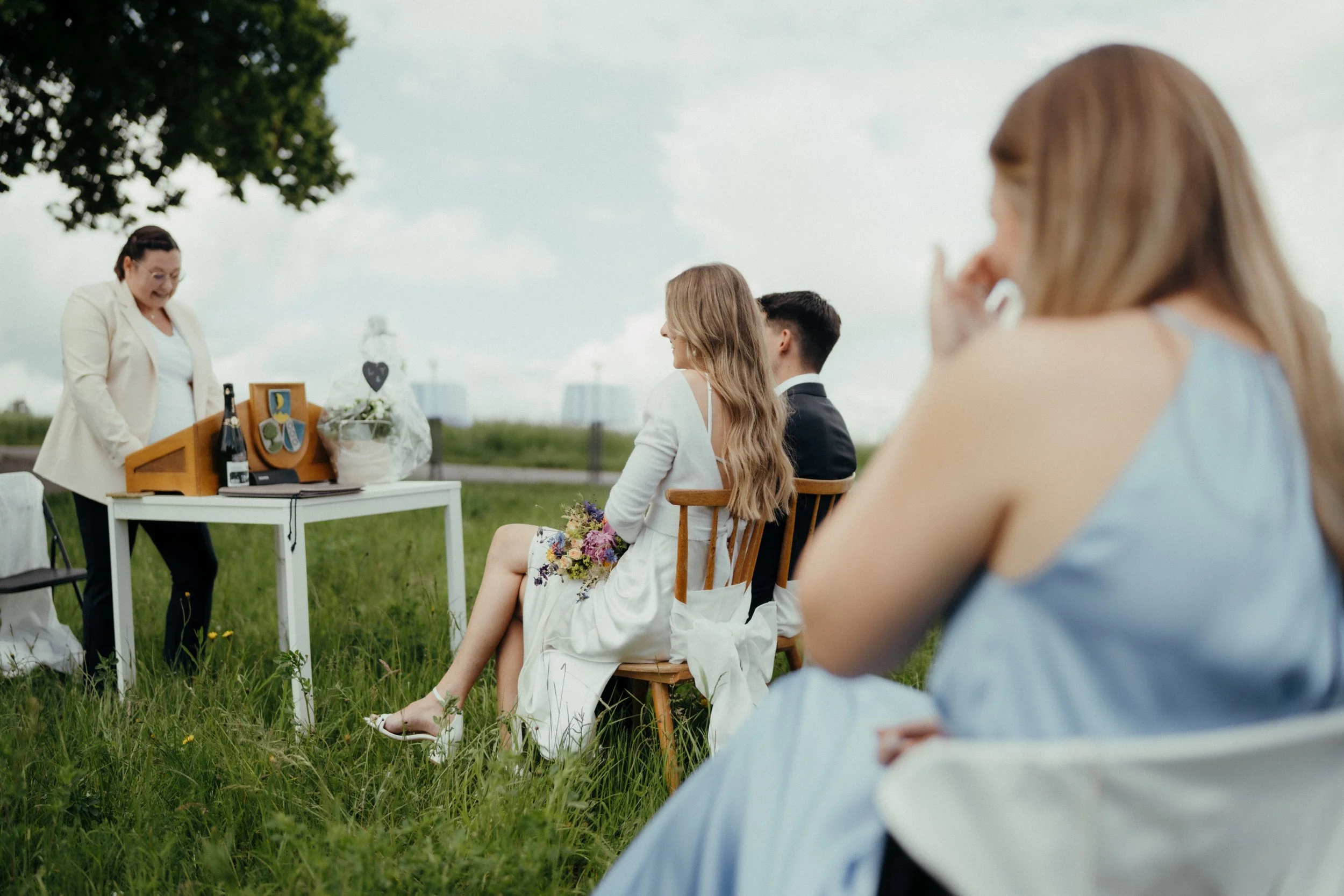 Bei einer Hochzeit im Freien stehen der Bräutigam und die Braut nebeneinander, während eine Frau mit weißen Jacke vor einem Tisch spricht. Es sitzen mehrere Gäste auf Stühlen, darunter eine Frau mit langem Brautkleid und Blumen im Schoß. Es ist bewöl