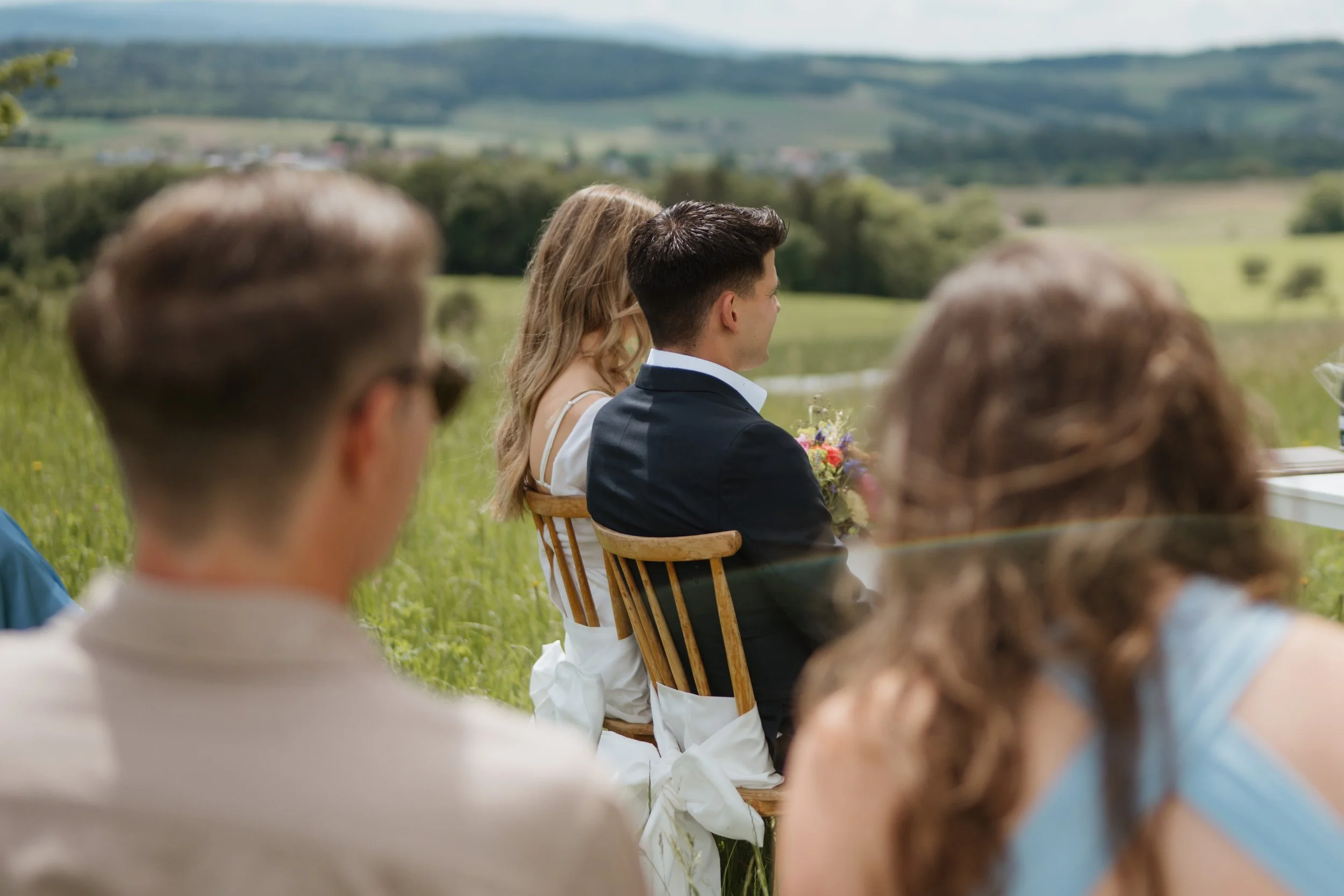 Eine Hochzeit im Freien mit mehreren Gästen, die in natürlicher Umgebung sitzen, im Hintergrund grüne Hügel und ein blauer Himmel.