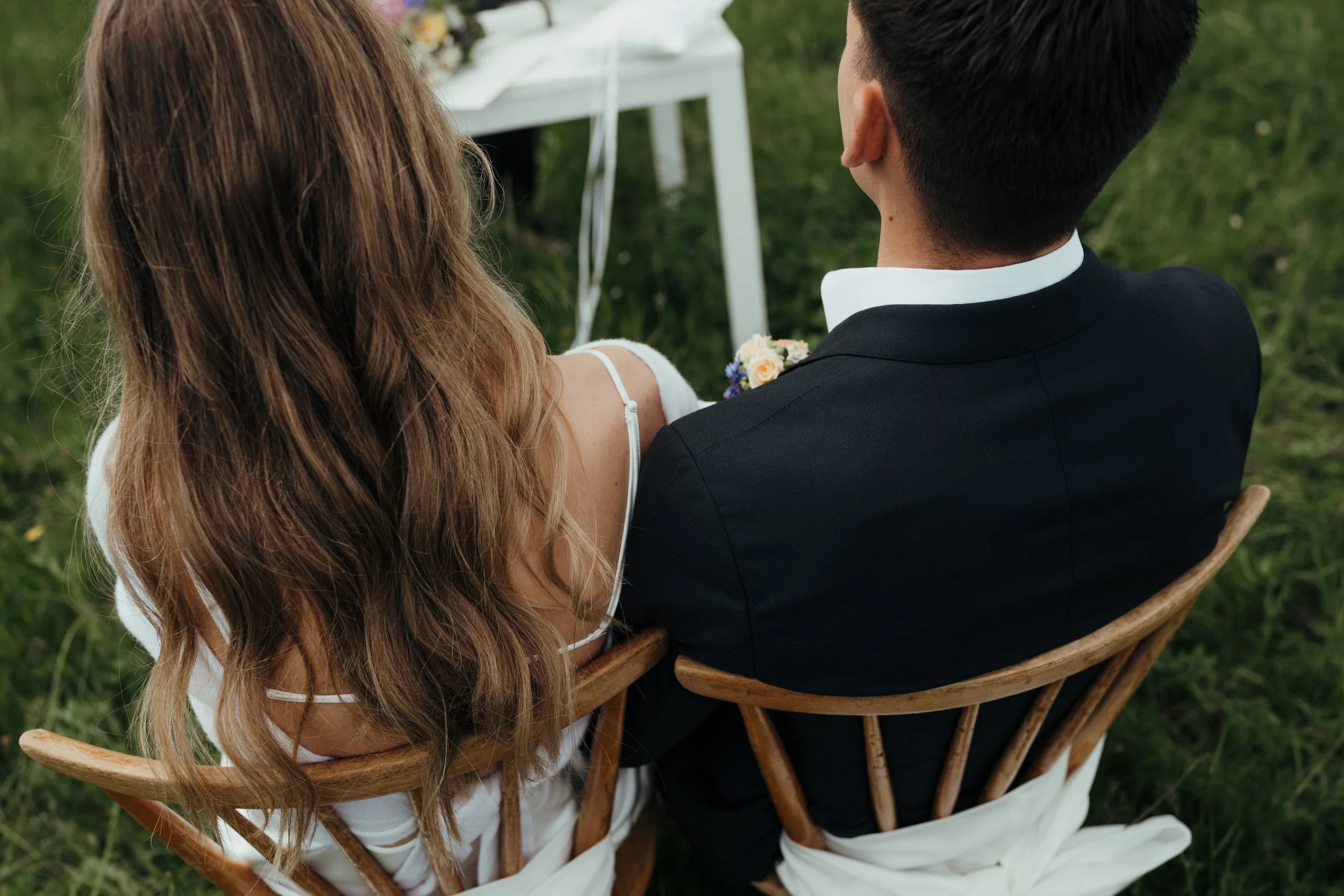 Ein Brautpaar sitzt auf einem Holzstuhl im Freien, während eine Hochzeit stattfindet. Die Frau trägt ein weißes Brautkleid mit dünnen Trägern, und der Mann trägt einen schwarzen Anzug. Im Hintergrund ist ein Baum und ein weißer Tisch zu sehen.