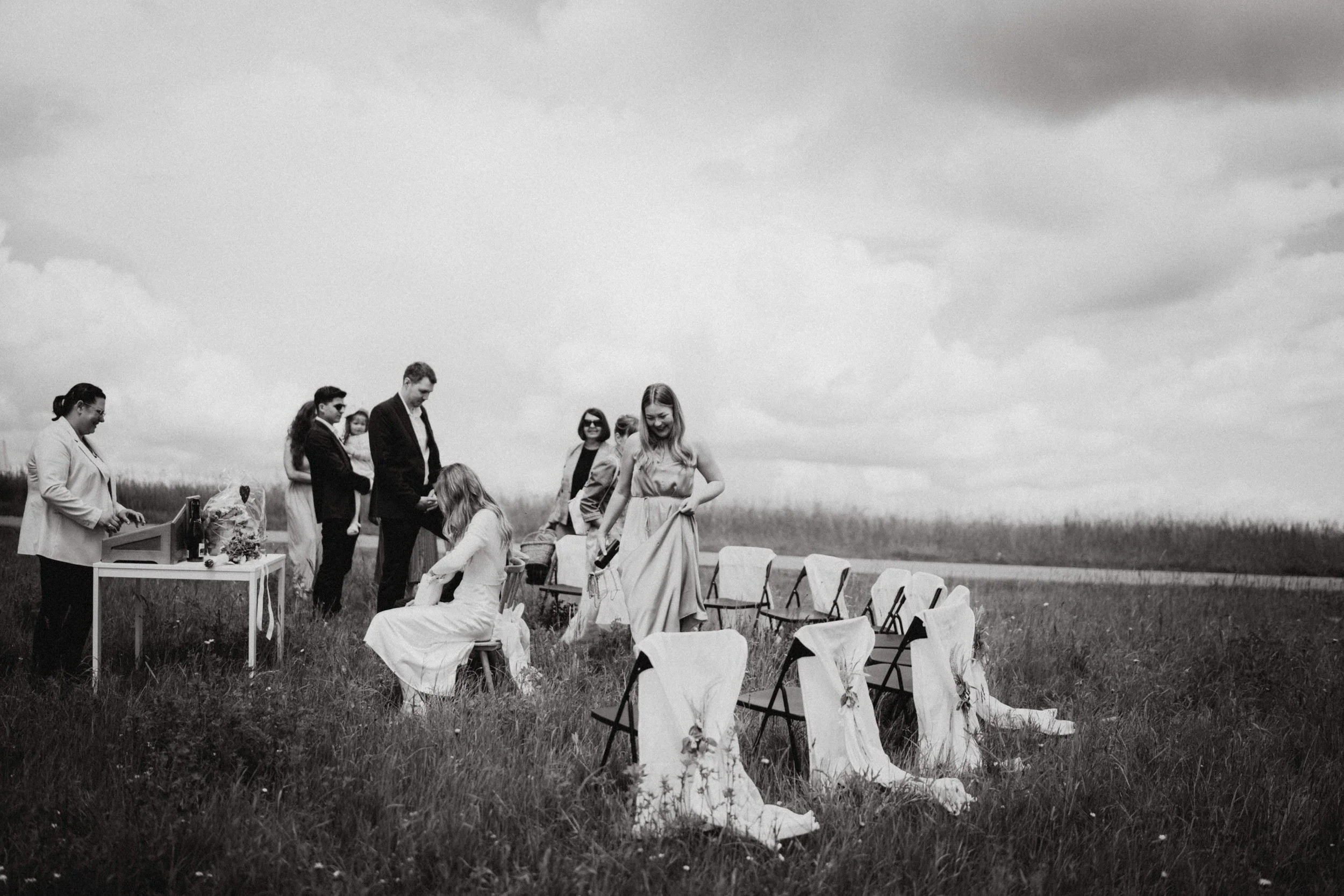 Eine Gruppe von Menschen, vermutlich bei einer Hochzeit im Freien, steht auf einer Wiese. Es sind Frauen und Männer, einige sitzen und andere stehen, alle lachen und scheinen fröhlich zu sein. Es gibt Stühle und Dekorationen, die auf eine Feier hinwe