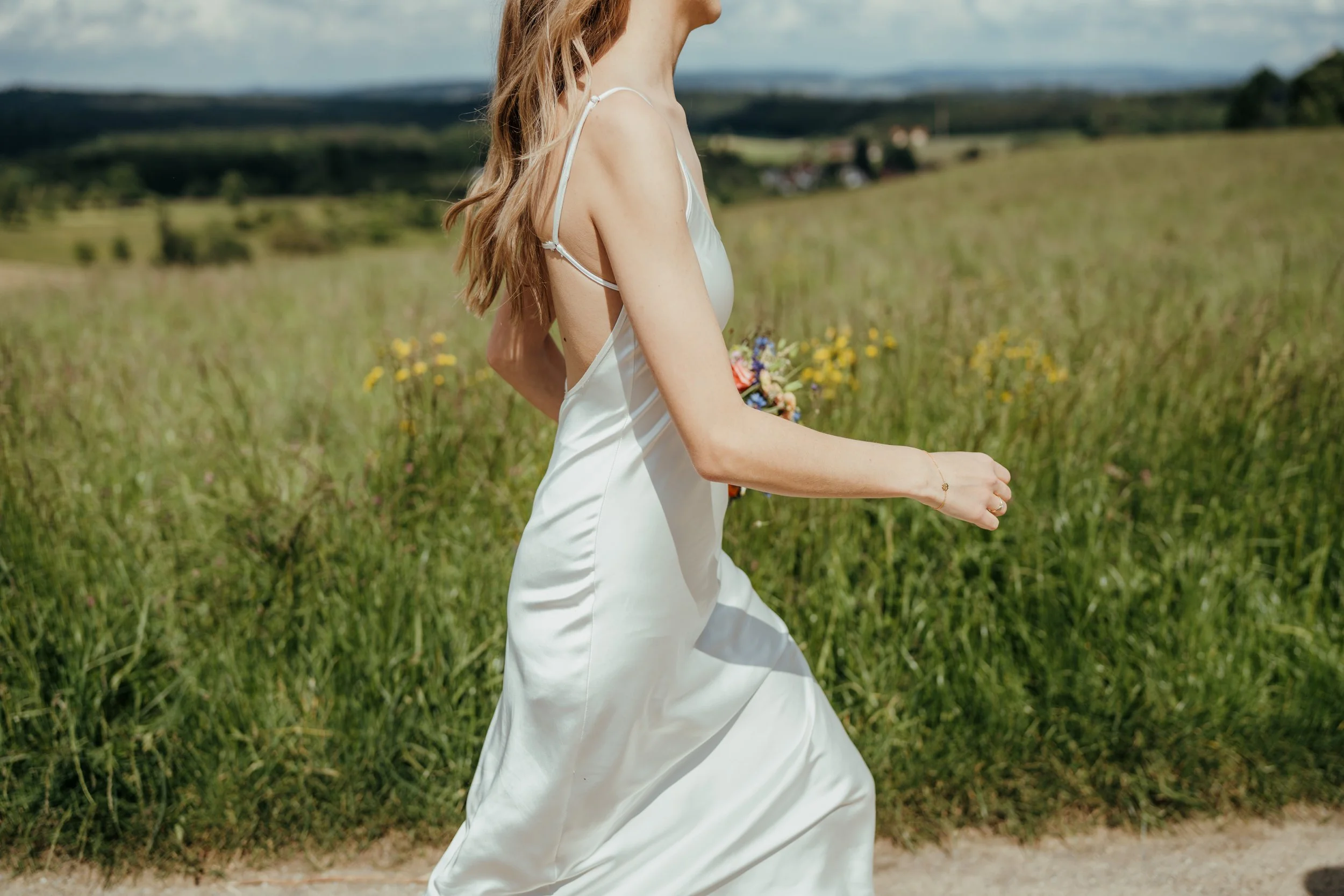 Frau in einem weißen Kleid, die durch eine grüne Wiesenlandschaft läuft.