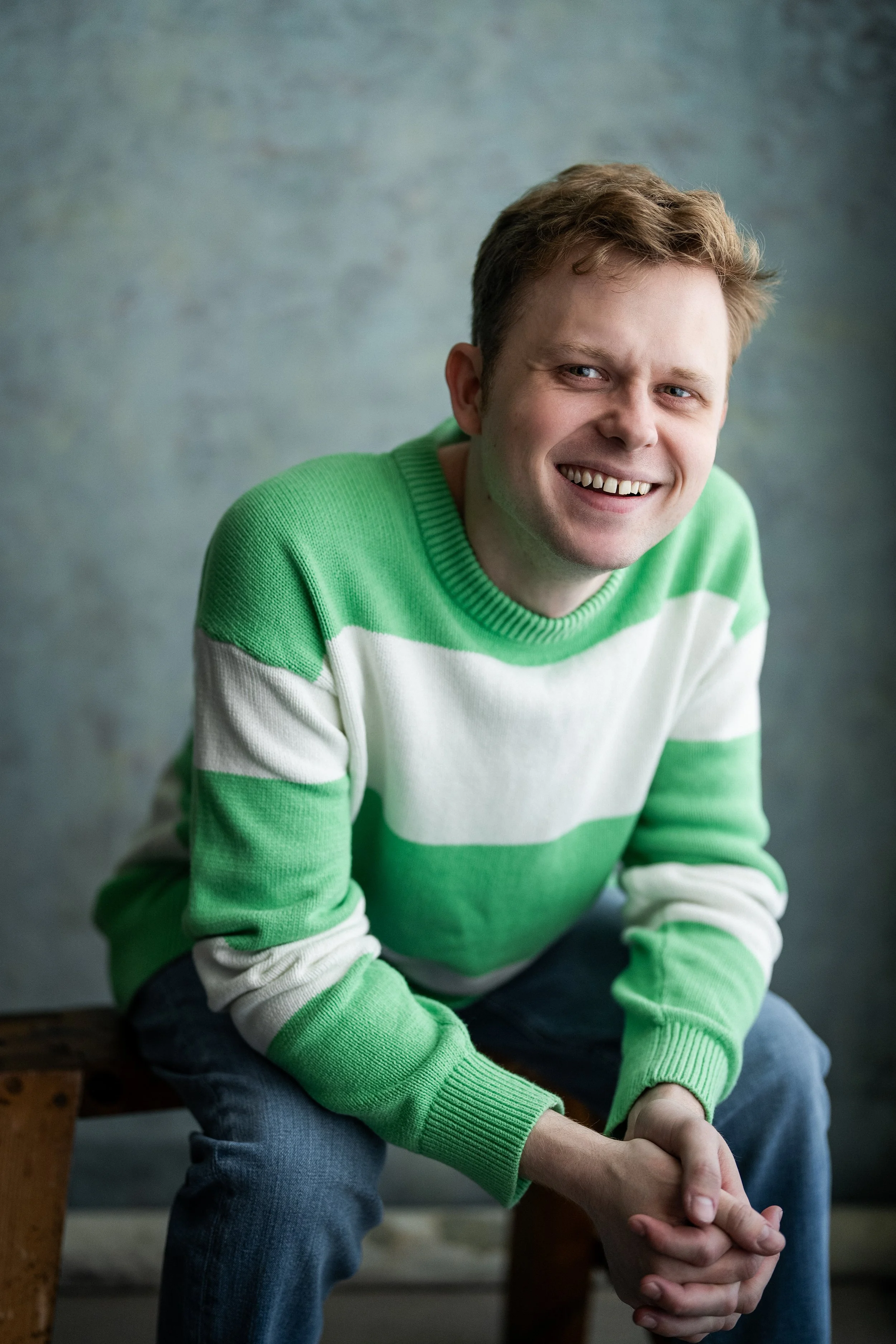 Man wearing a green and white striped sweater, smiling and sitting on a wooden stool against a textured gray-blue background.