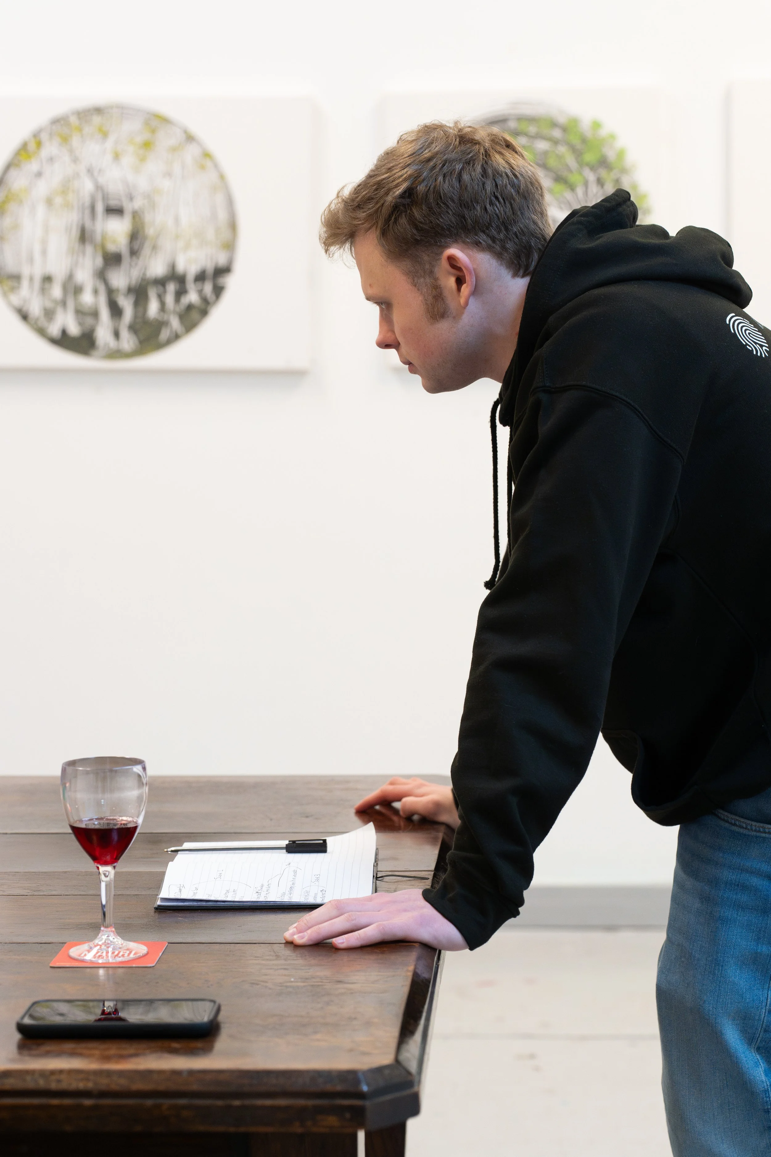 A person in a black hoodie is leaning over a wooden table. On the table, there is a glass of red wine, a notepad with a pen, and a smartphone. Artwork is visible on the wall in the background.