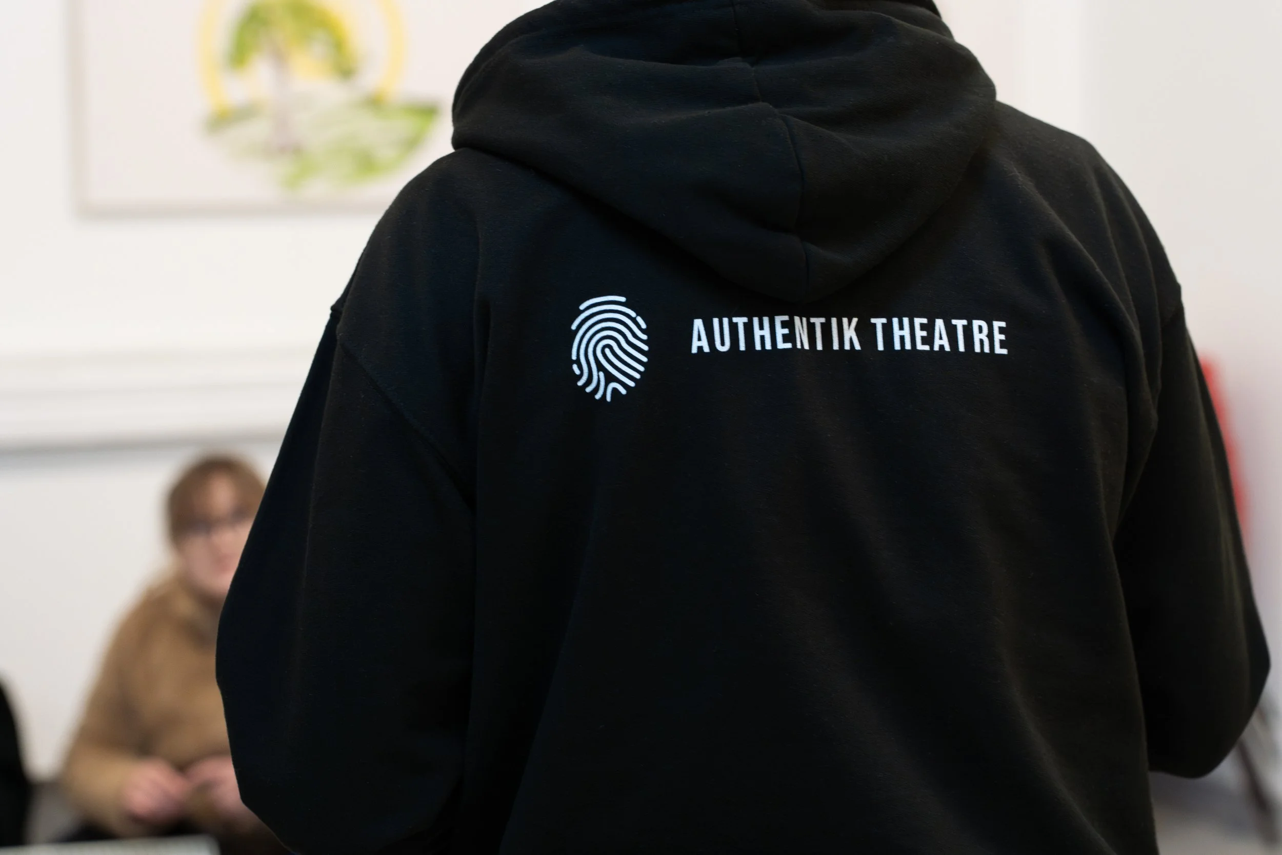 Person wearing a black hoodie with 'Authentik Theatre' and a fingerprint logo on the back, sitting in a room with a blurred person in the background.