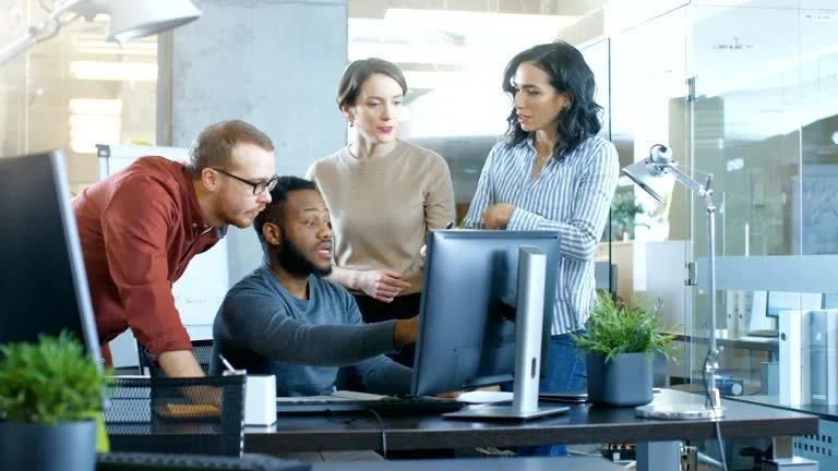 Four diverse professionals gathered around a computer in a modern office, discussing work.