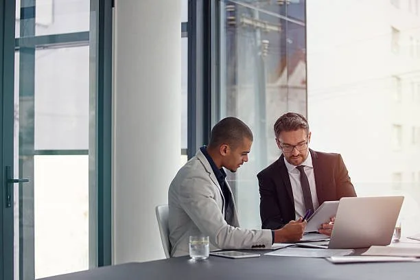Two business professionals, one man and one woman, sitting at a table in an office, reviewing documents on a laptop with a glass of water nearby.