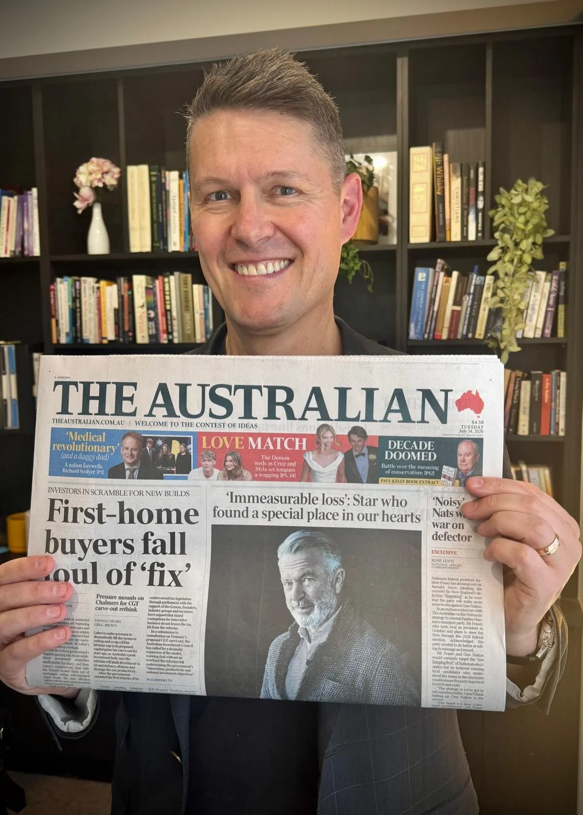 A smiling Andre Knott holding a copy of The Australian newspaper in front of a bookshelf, with books and a flower vase visible in the background.