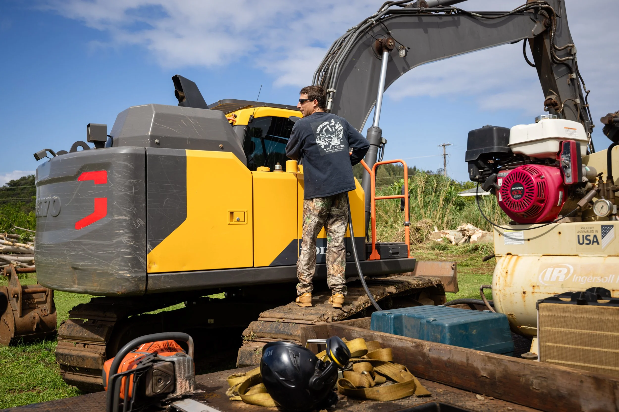 Man refueling a yellow excavator while standing on tracks, with a construction site setting and various equipment visible around.