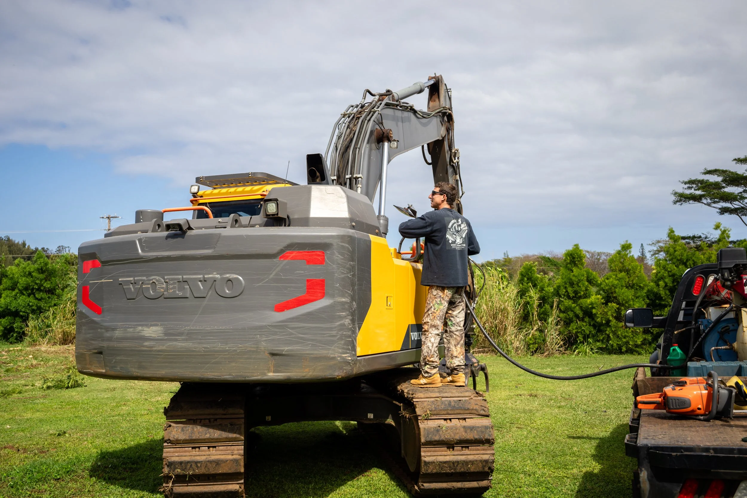 Person fueling a Volvo excavator on grass near trees under a cloudy sky.