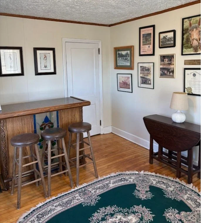 Corner of a room with a wooden bar counter, three wooden bar stools, a small side table with a lamp, framed pictures on the walls, and a green floral patterned rug on a hardwood floor.