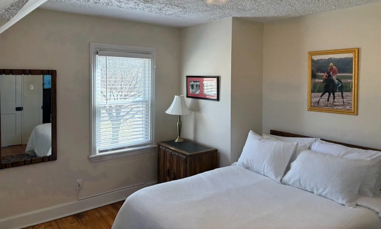 A bedroom with a bed, a window with blinds, a mirror, a nightstand with a lamp, and pictures on the wall, including a framed photo of a horse race.
