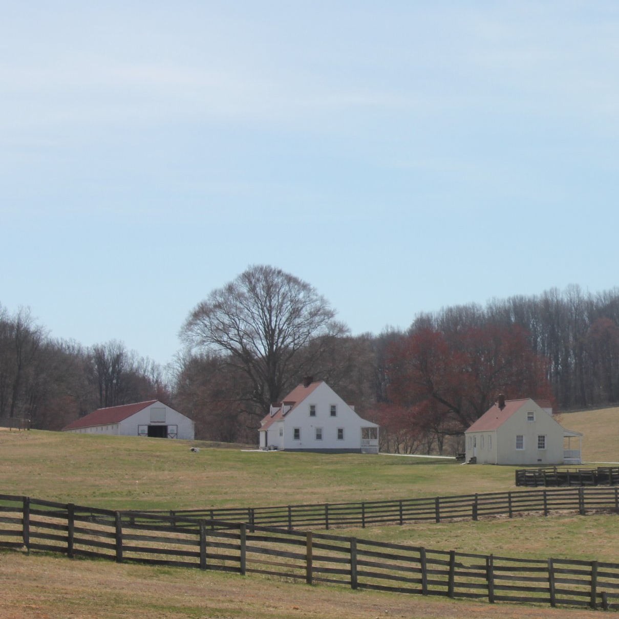 A rural farm scene with white houses, barn, and wooden fences, surrounded by leafless trees on a cloudy day.