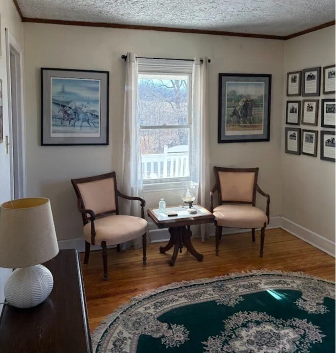 A small room with two armchairs, a side table, a framed picture, a wall of photographs, a window with curtains, a ceiling light, and a round patterned rug.