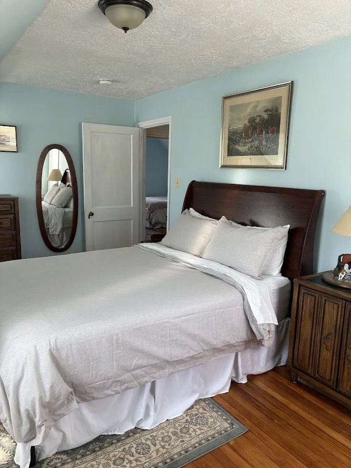 A cozy bedroom with a large bed, wooden headboard, and light blue walls. There is a framed picture above the bed, a lamp on a wooden nightstand, and a mirror reflecting another bed in an adjacent room.
