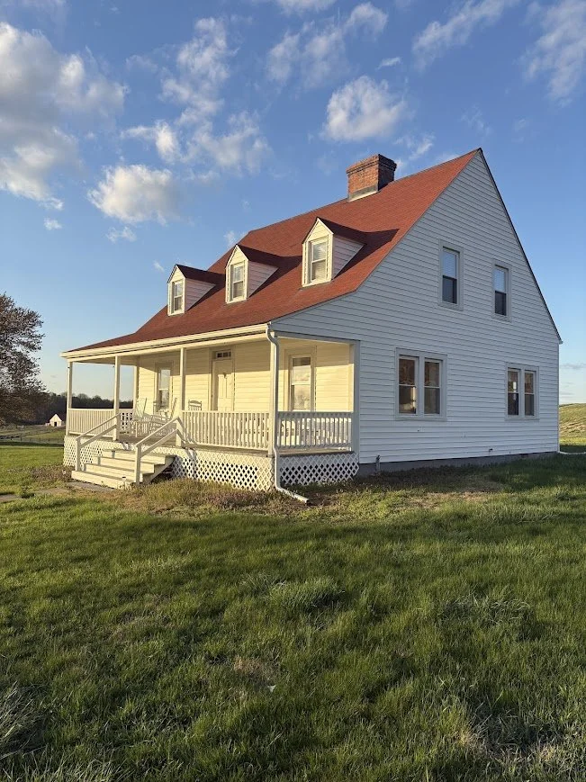 A white two-story house with a red gabled roof, three front dormer windows, a front porch, and a brick chimney, set on a grassy landscape under a partly cloudy sky.