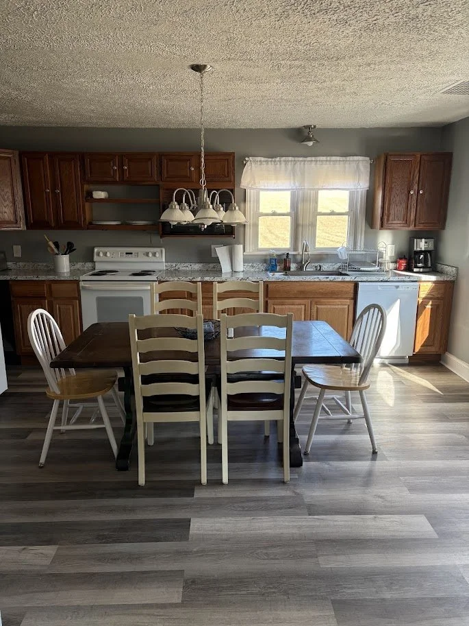 A kitchen with wooden cabinets, a wooden dining table with six chairs, a window above the sink, and a light fixture hanging above the table.