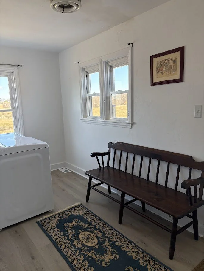 A laundry room with a white washing machine, a wooden bench, a small patterned rug, and two windows with views of a grassy field and blue sky.
