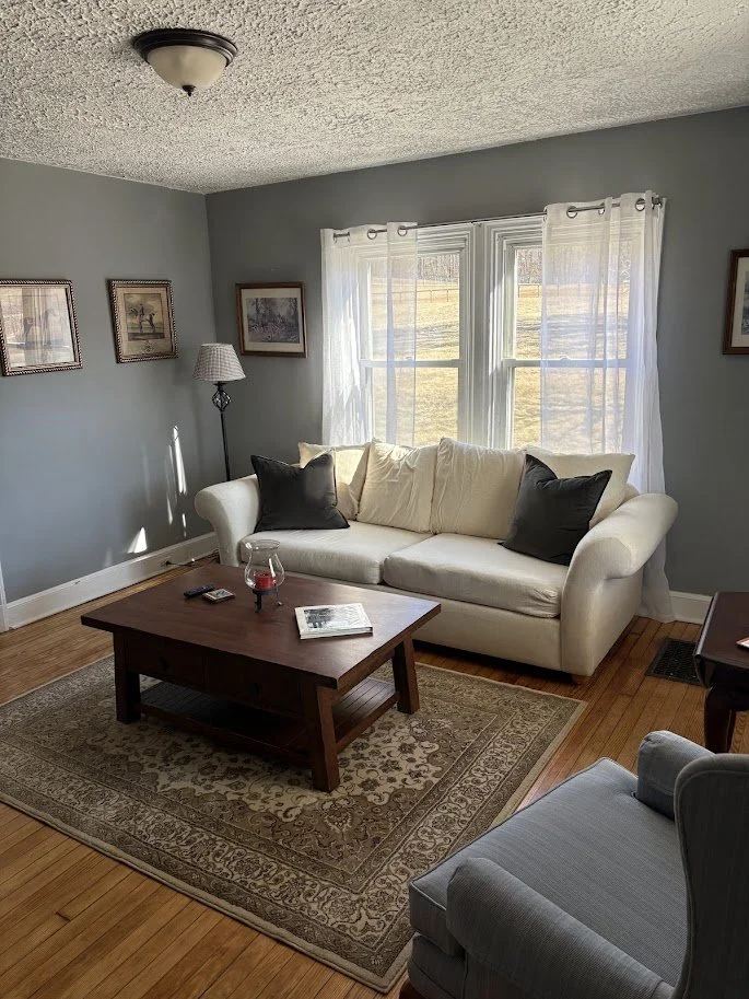 Living room with a white sofa, black and beige pillows, wooden coffee table, area rug, window with white curtains, gray wall, framed pictures, floor lamp, and hardwood floor.