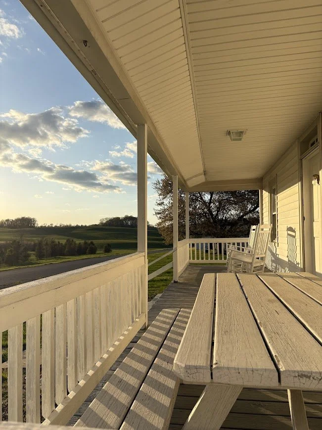 Sunset view from a porch with a wooden table and white rocking chairs, overlooking a grassy landscape with trees and a partly cloudy sky.