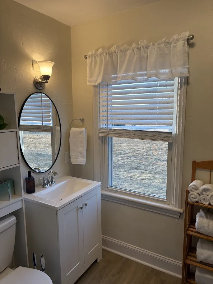 Small bathroom with a white vanity, oval mirror, wall-mounted light fixture, window with white blinds and ruffled valance, towel hanging on wall, and shelves with folded towels.