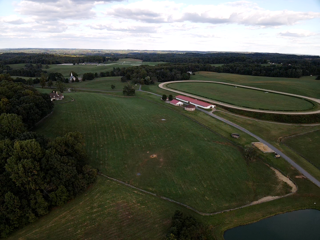 An aerial view of a bonita farm in darlington maryland with green fields, a horse racetrack, several farm buildings, trees, a driveway, and a pond in the foreground under a partly cloudy sky.