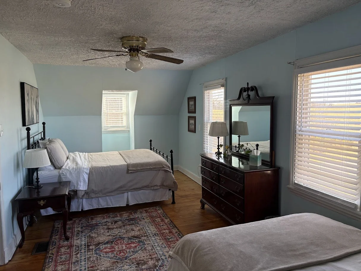 A bedroom with two beds, wooden furniture, a ceiling fan, and multiple windows with blinds.