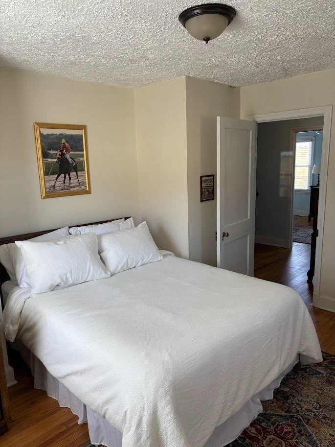 A bedroom with a bed covered in white bedding and pillows, a framed picture of a person riding a horse on the wall, and an open door leading to a hallway with hardwood floors and a window.