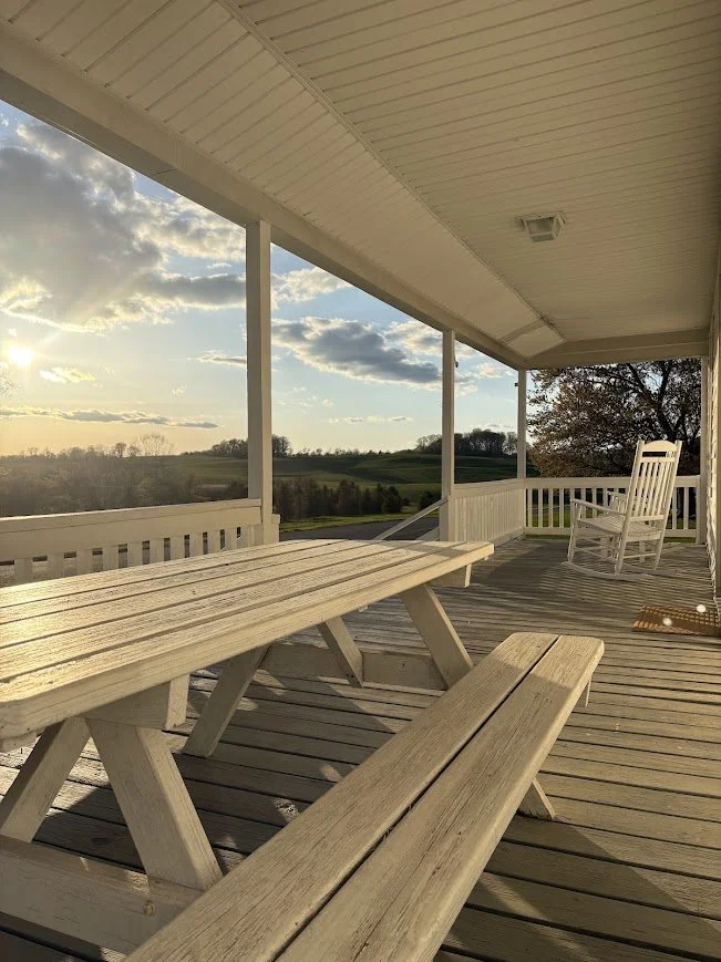 A wooden porch with a picnic table and a rocking chair, overlooking a green landscape with trees and a cloudy sky at sunset.
