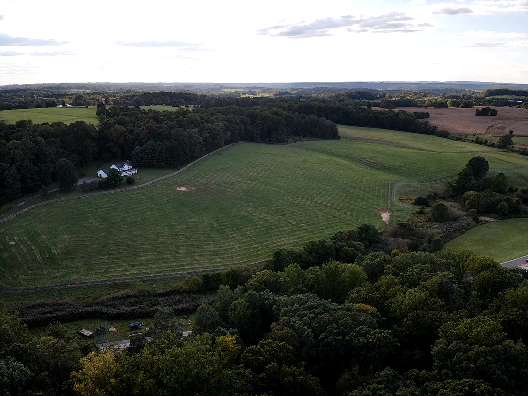 Aerial view of a rural landscape with open fields, a few houses, and dense tree cover, under a partly cloudy sky.