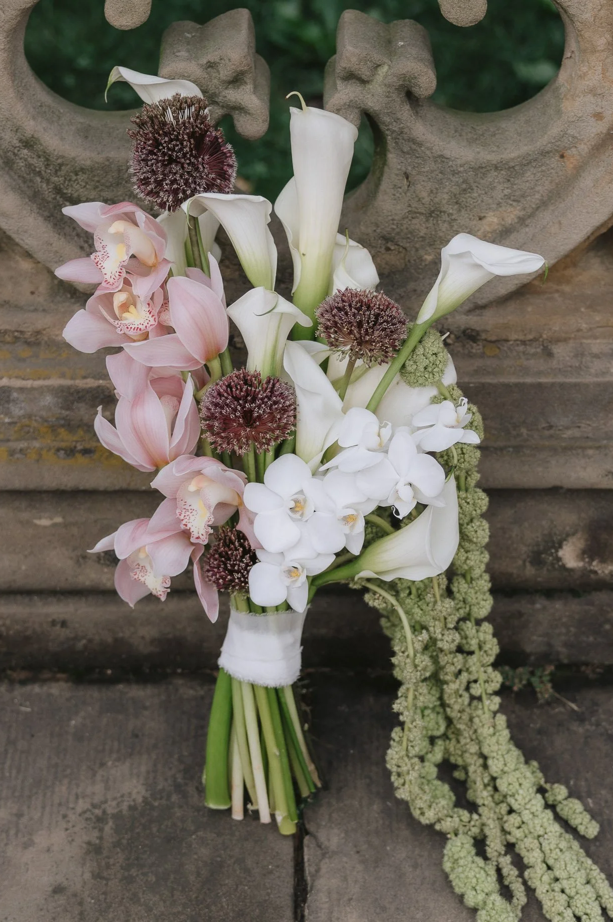 A bridal bouquet of pink cymbidium orchids, white calla lilies, and allium flowers, with a trailing green amaranth flower