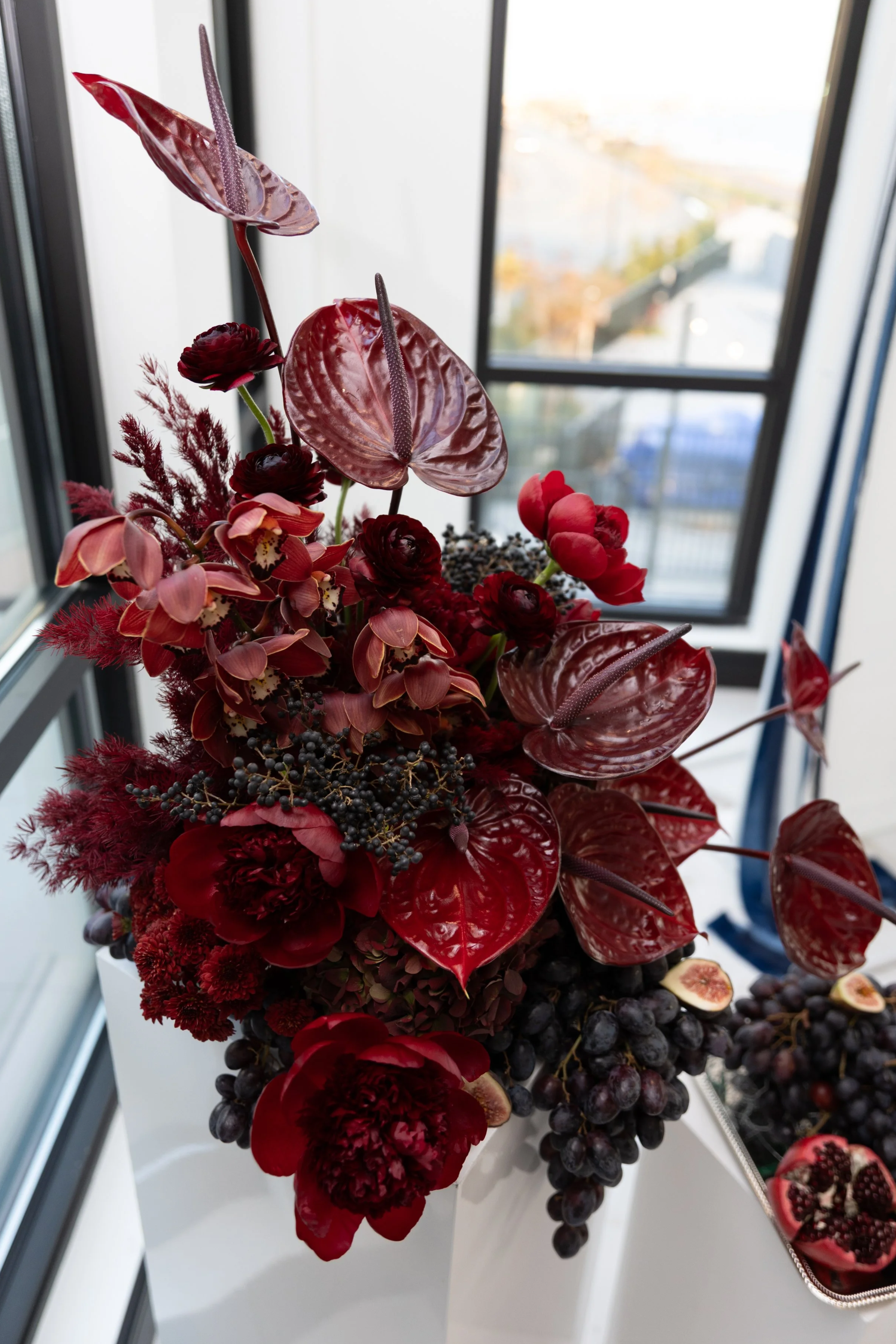 Stunning modern floral arrangement featuring burgundy and deep red flowers such as hydrangea, anthurium, mum flower, cymbidium orchid; trailing dark grapes