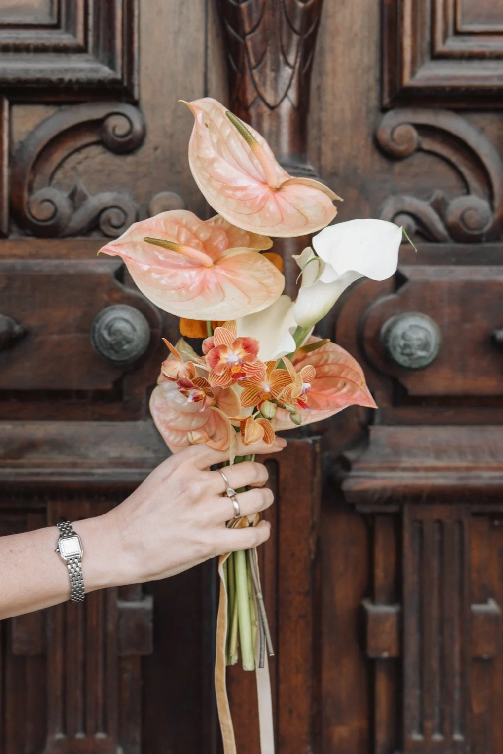  Photo of a bride holding a modern bouquet of peach and white flowers, including anthuriums, orchids and callas