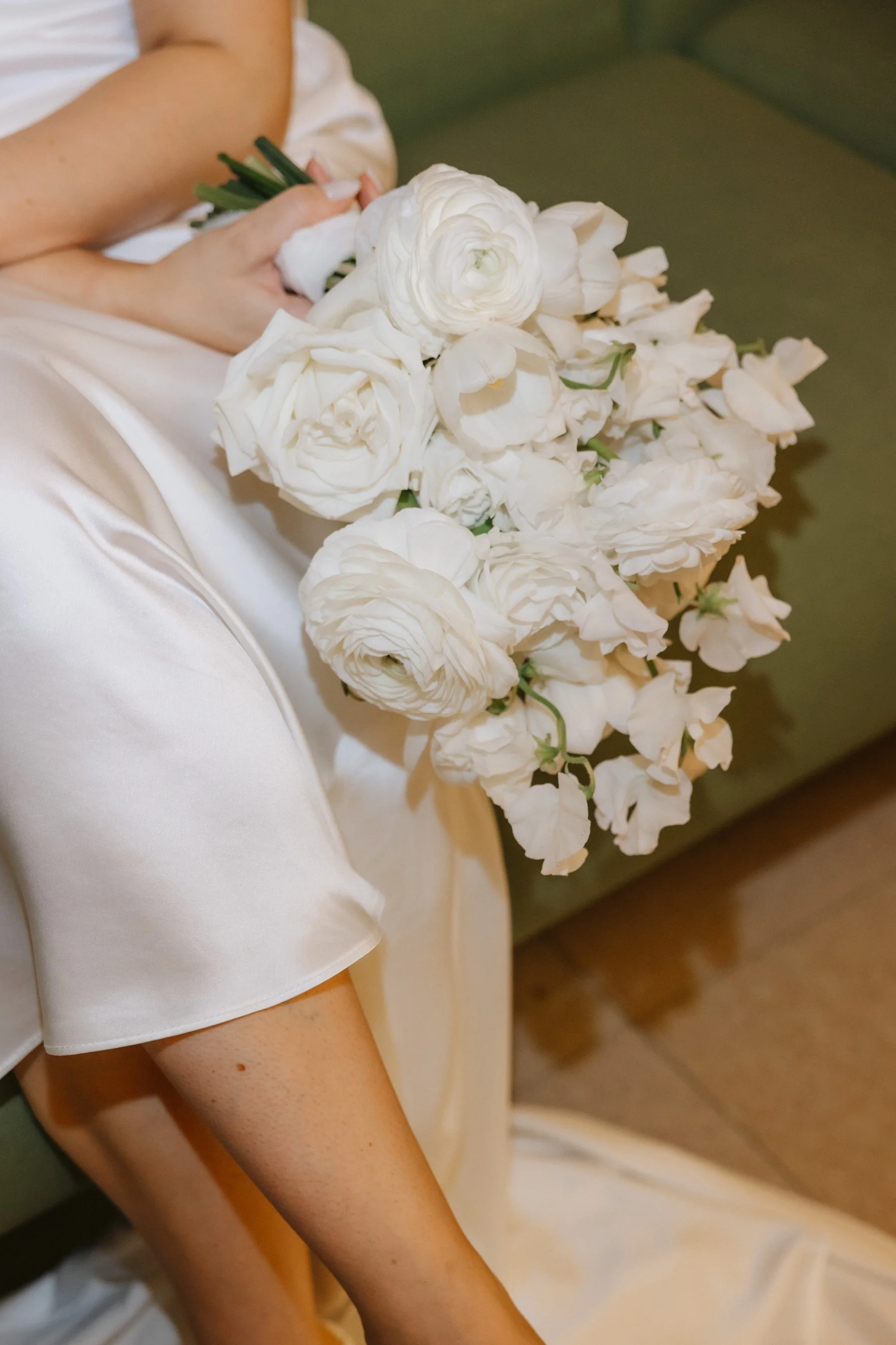 A bride in a white satin dress holding a airy bridal bouquet of white flowers: sweet pea, ranunculus, roses, tulips