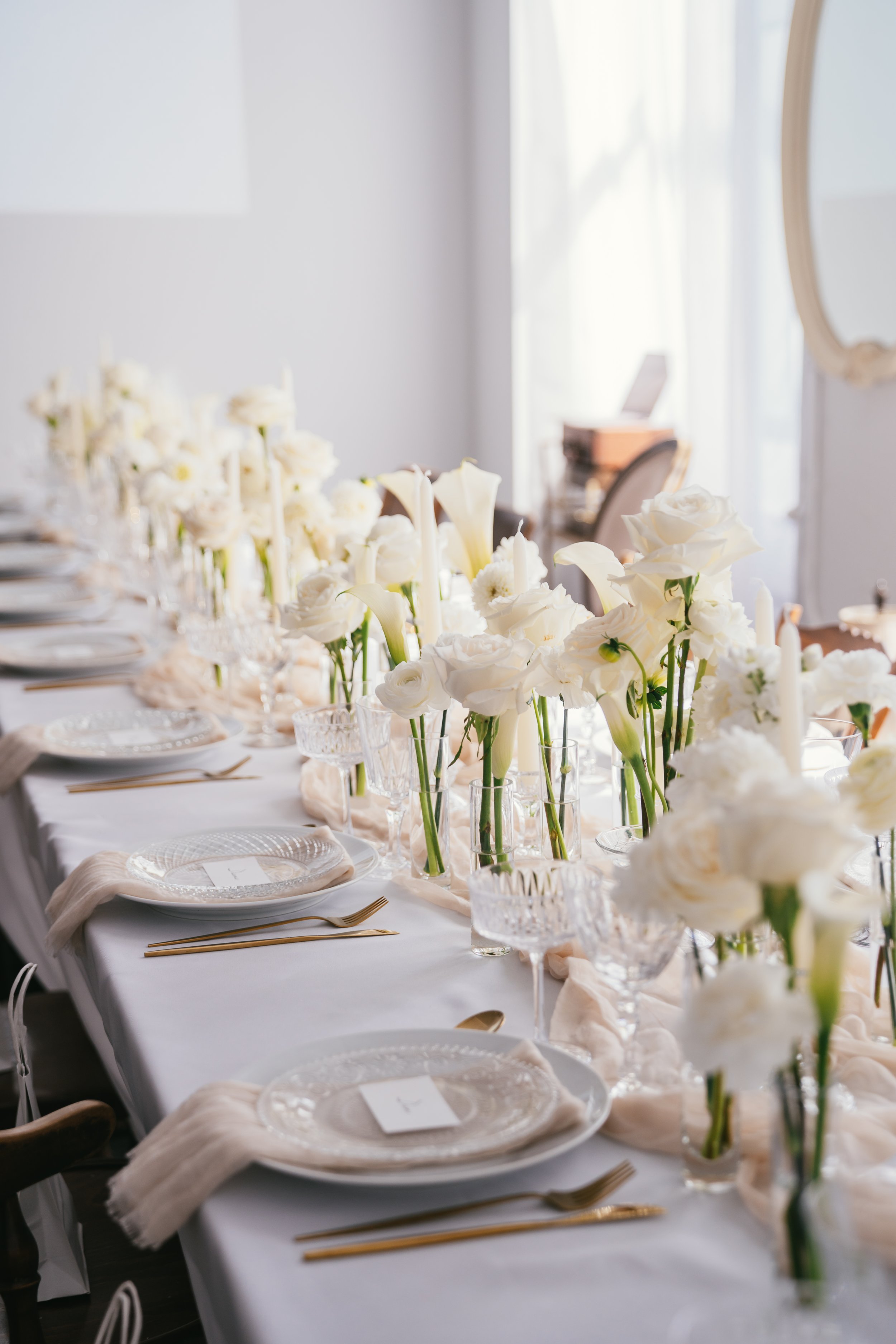 Elegant wedding reception table decorated with bud vases with white flowers: calla lily, roses, ranunculus, stock flower