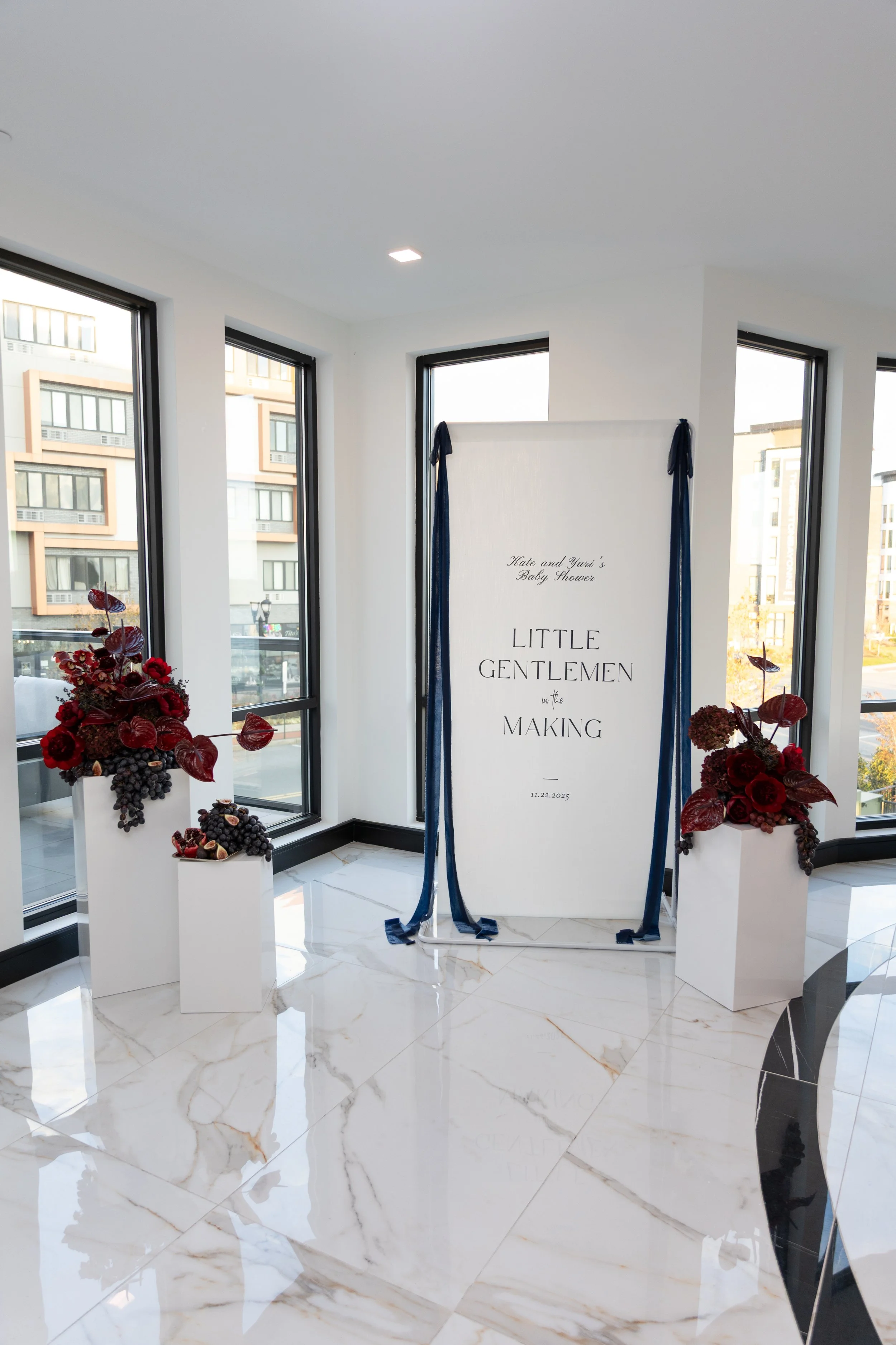 A baby shower display with a sign saying 'Little Gentlemen in the Making', decorated with dark blue ribbons and surrounded by sculptural and whimsical floral arrangements with burgundy and dark red flowers and grapes