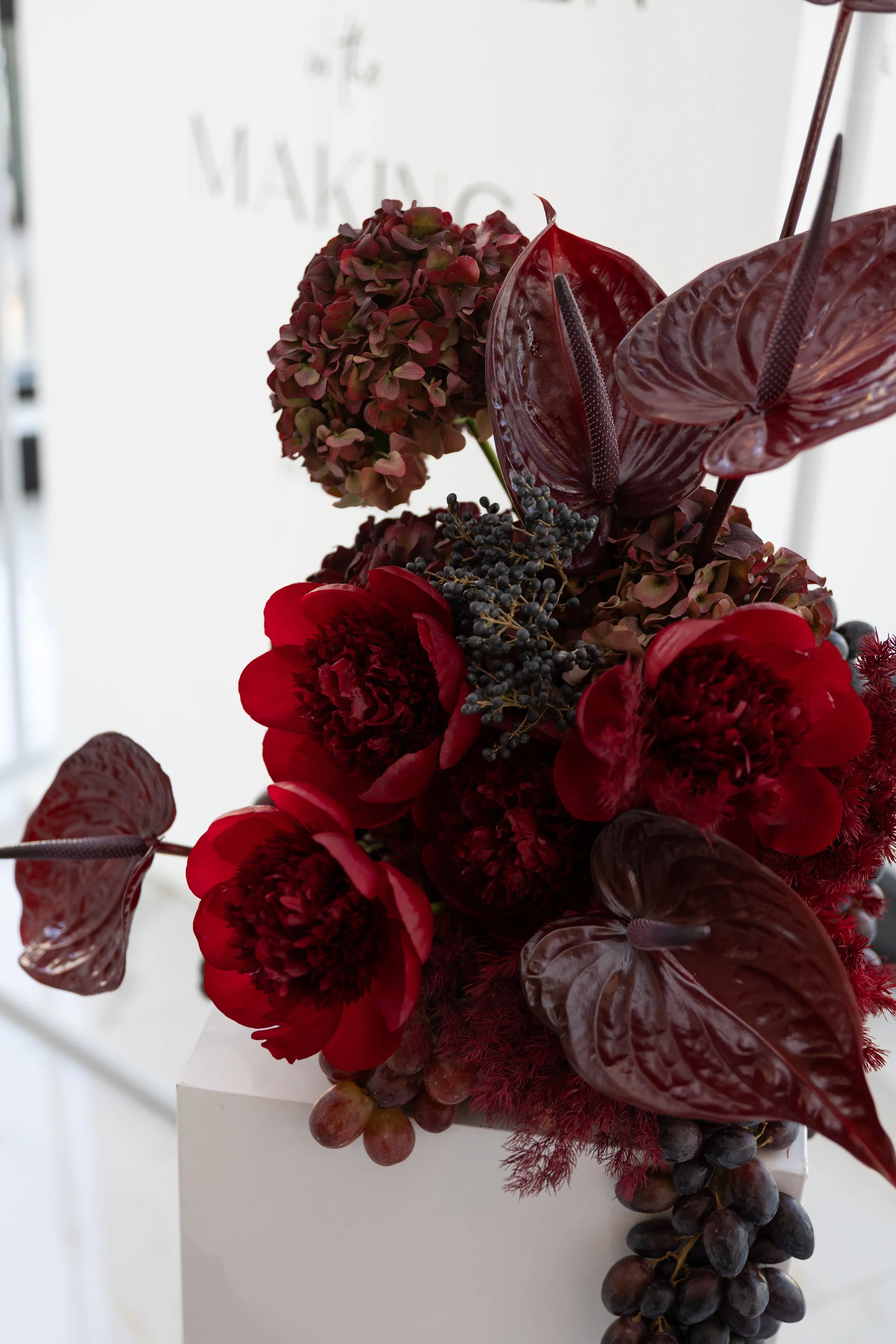 A sculptural floral arrangement with burgundy peonies, dark red anthuriums, premium hydrangea and a cluster of hanging grapes