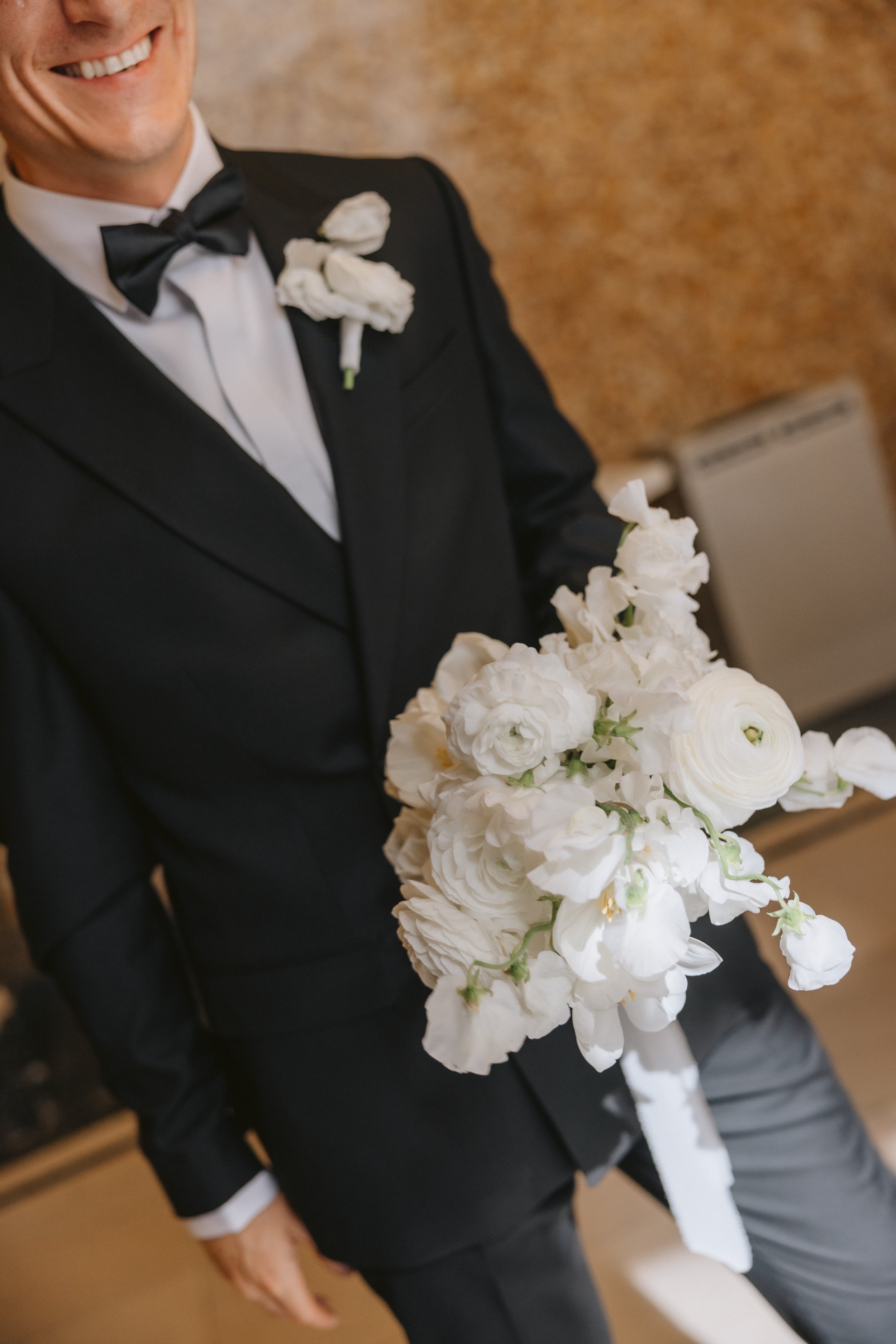 A groom dressed in a black tuxedo with a bow tie, holding a modern bouquet of white flowers such as sweet pea, ranunculus, tulips and roses