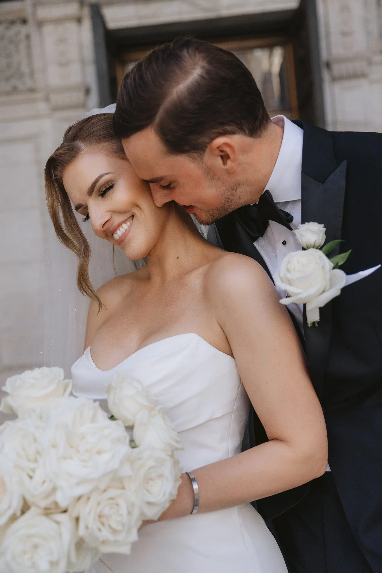 A bride and groom on their wedding day holding gorgeous bridal bouquet of white roses. Groom is wearing stunning boutonniere