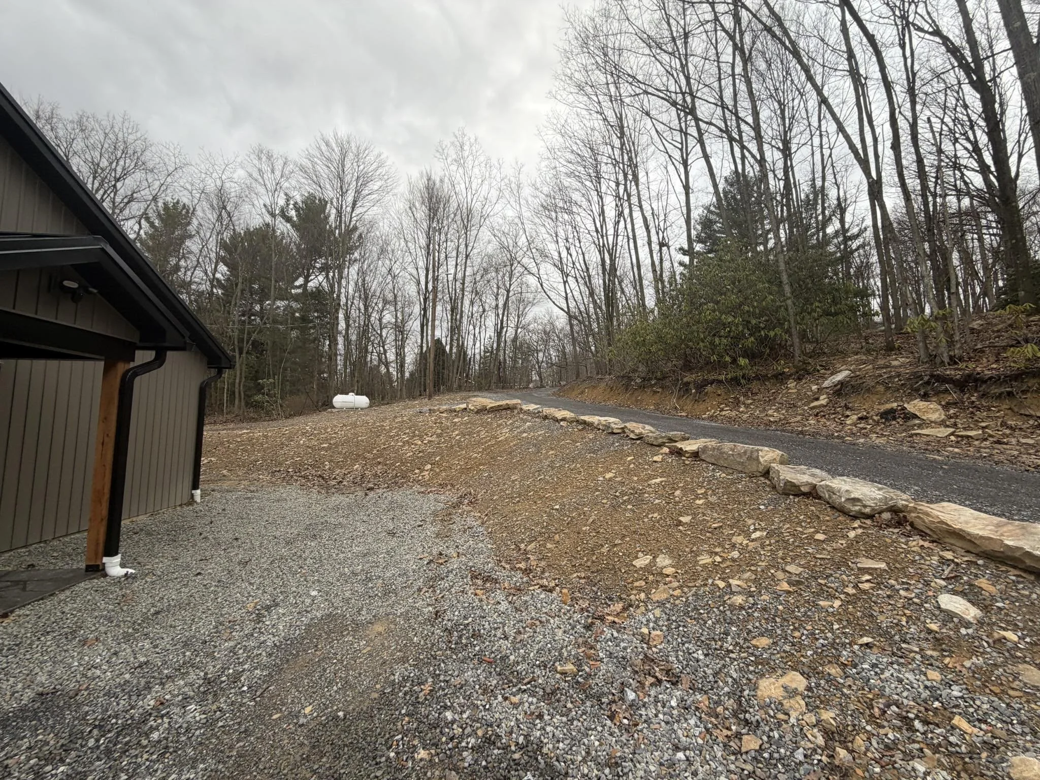 A gravel driveway with a stone border, a house partially visible on the left, and a wooded hillside with leafless trees in the background. Overcast sky.