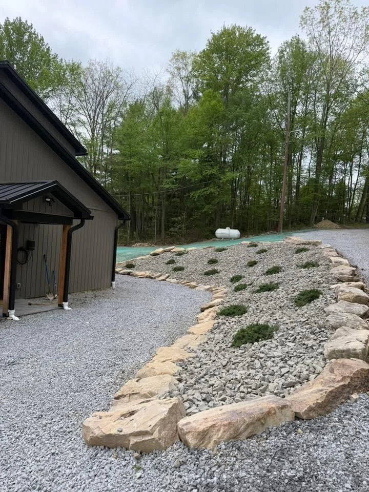 Residential backyard with gravel pathway bordered by large rocks, gravel garden bed with small bushes, and a wooded area in the background