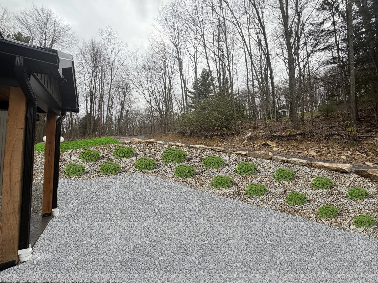 A backyard with a gravel patio, a stone retaining wall with small green shrubs, and a wooded hillside background with leafless trees.