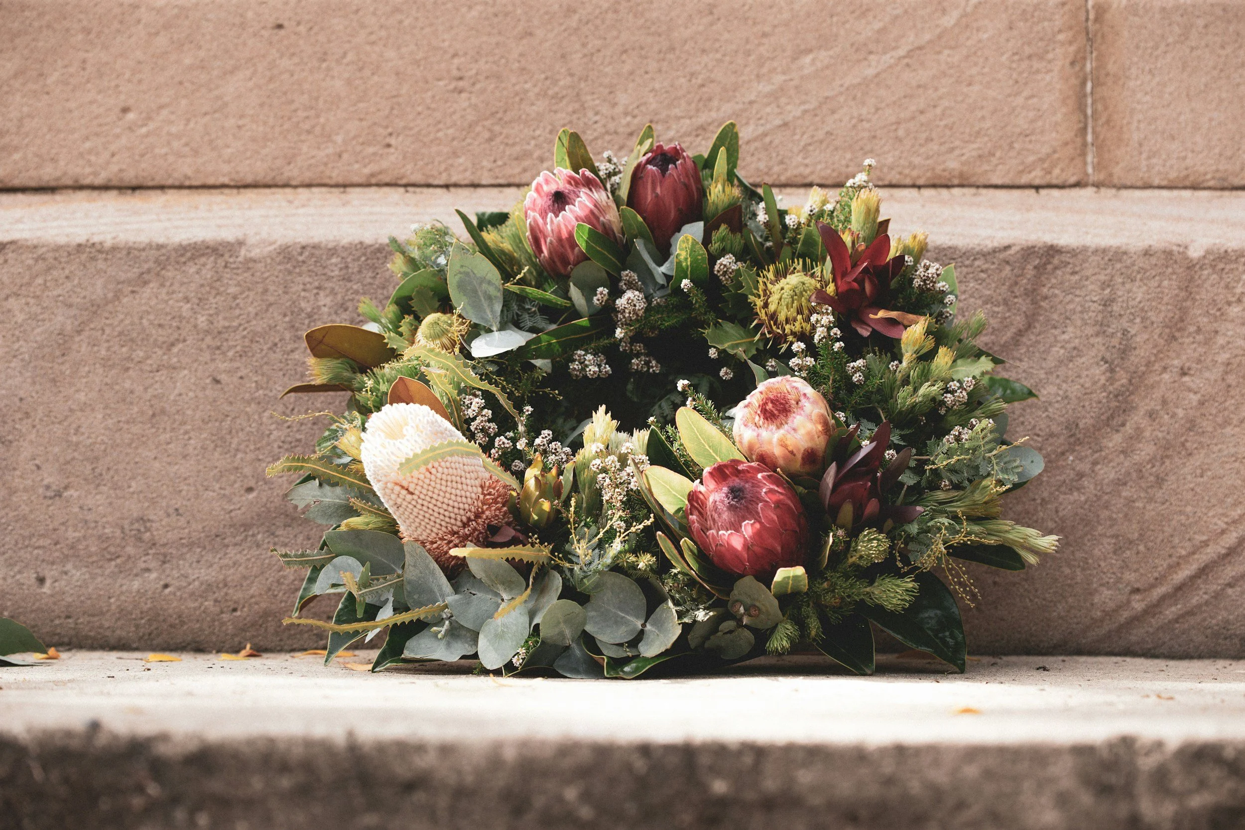 A floral arrangement with protea flowers, eucalyptus leaves, and small white filler flowers placed on a stone ledge in front of a stone wall.