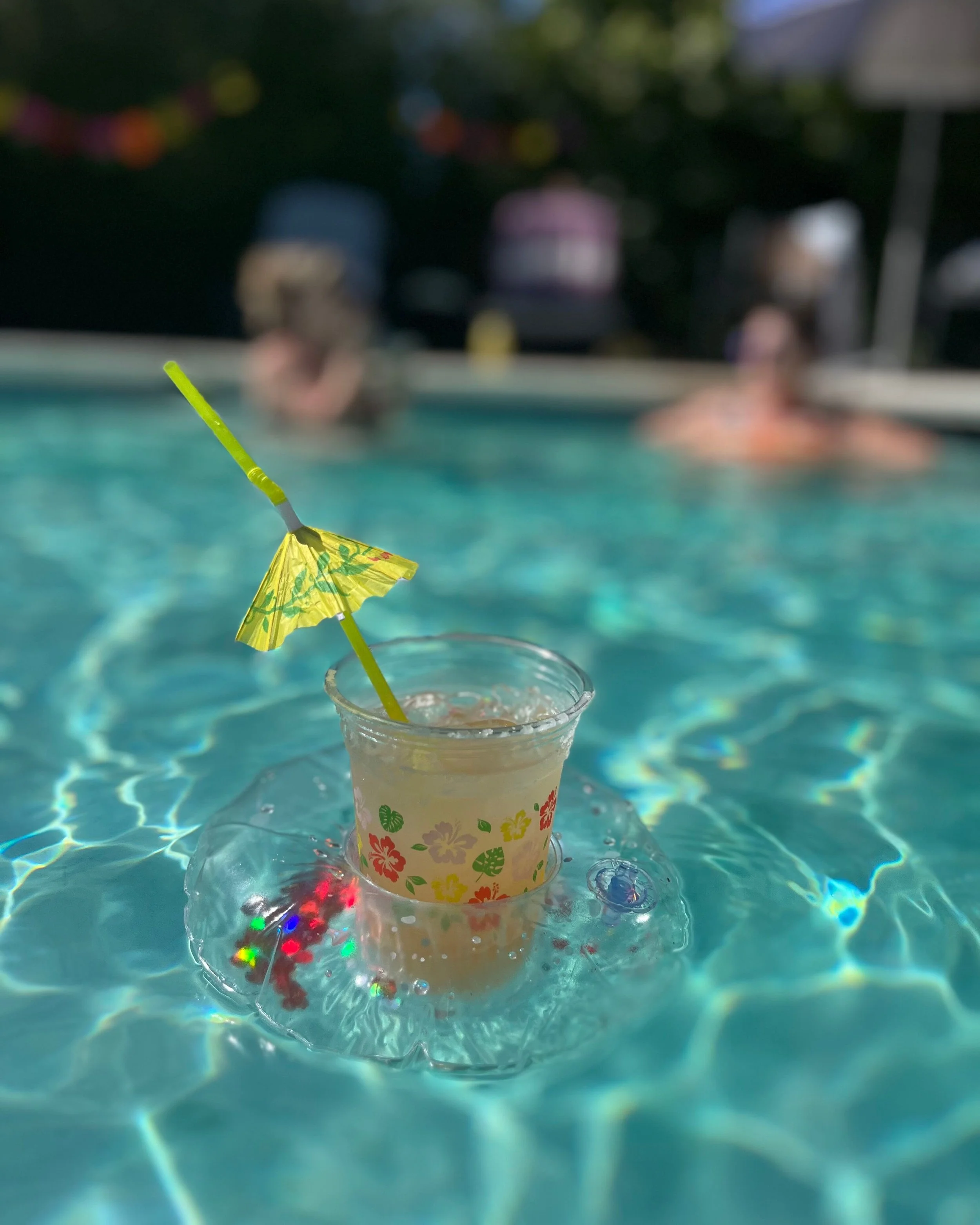 A tropical drink with a yellow umbrella and straw floating on a pool, with two people swimming in the background.