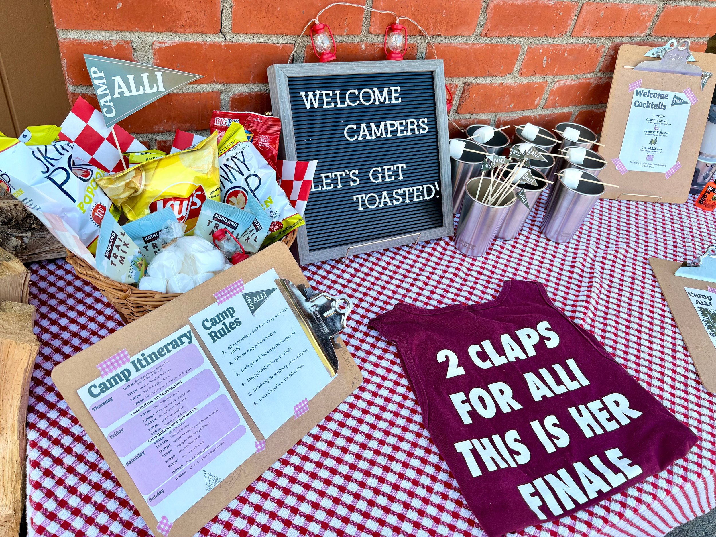 Camping-themed table setup with a sign that says "WELCOME CAMPERS. LET'S GET TOASTED!", snack bags, cups with straws, camp rules, a towel with "2 CLAPS FOR ALLI THIS IS HER FINAL" written on it, and a picnic tablecloth.