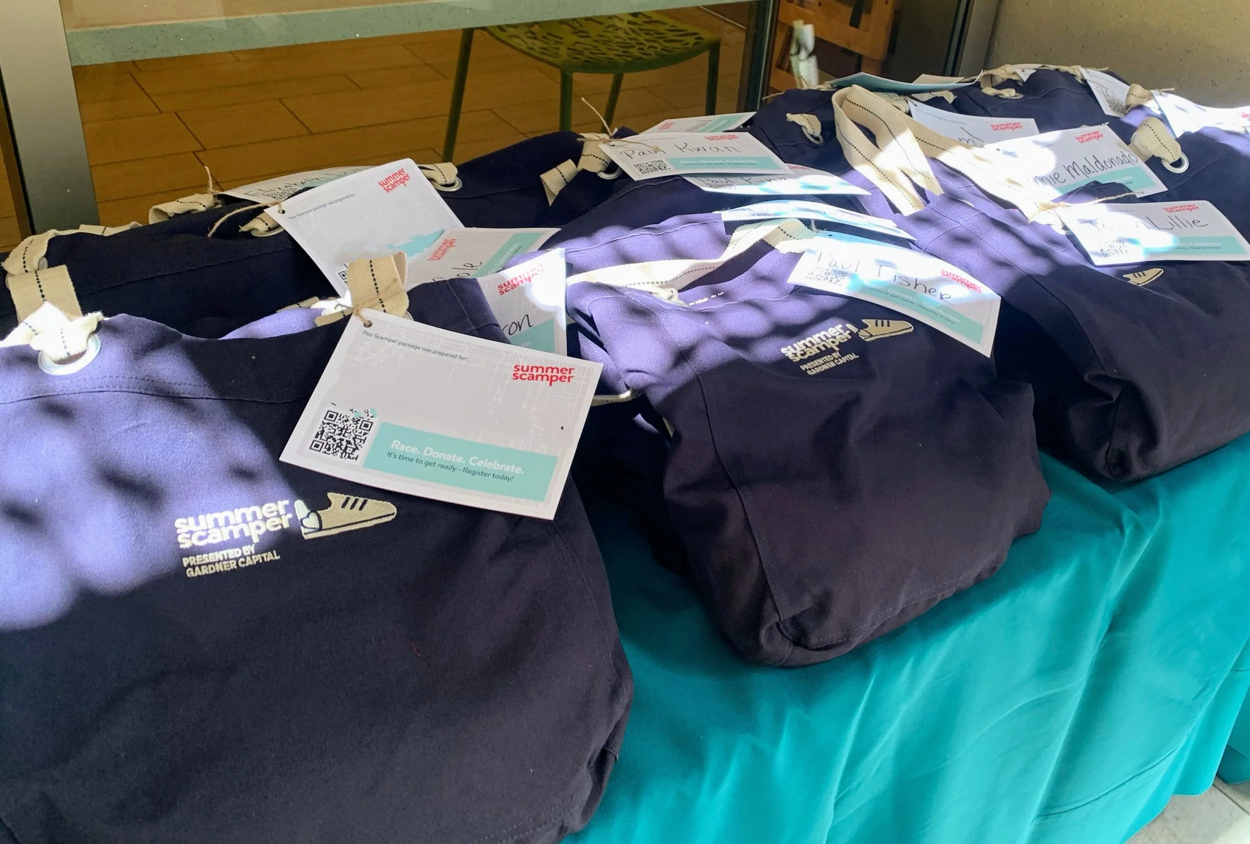 Several navy blue and purple tote bags on a teal cloth-covered table, each with a tag attached. The bags feature the Summer Scampers logo and are prepared for a summer event.