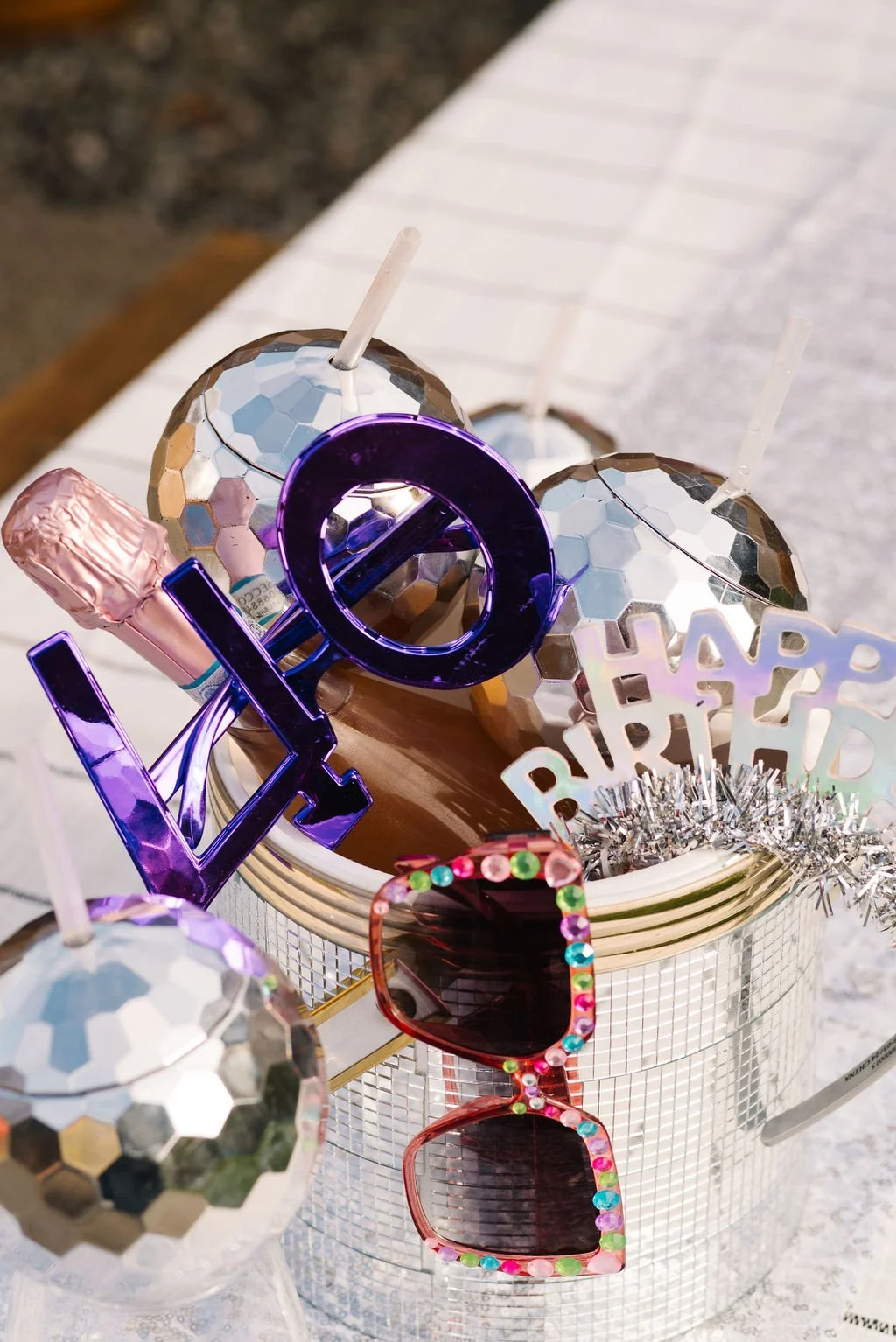 Decorations for a birthday celebration, including disco ball ornaments, a purple 'LOVE' sign, a 'HAPPY BIRTHDAY' cutout, tinsel, a pair of sunglasses with multicolored rhinestones, and a champagne bottle sculpture, all arranged on a white surface.