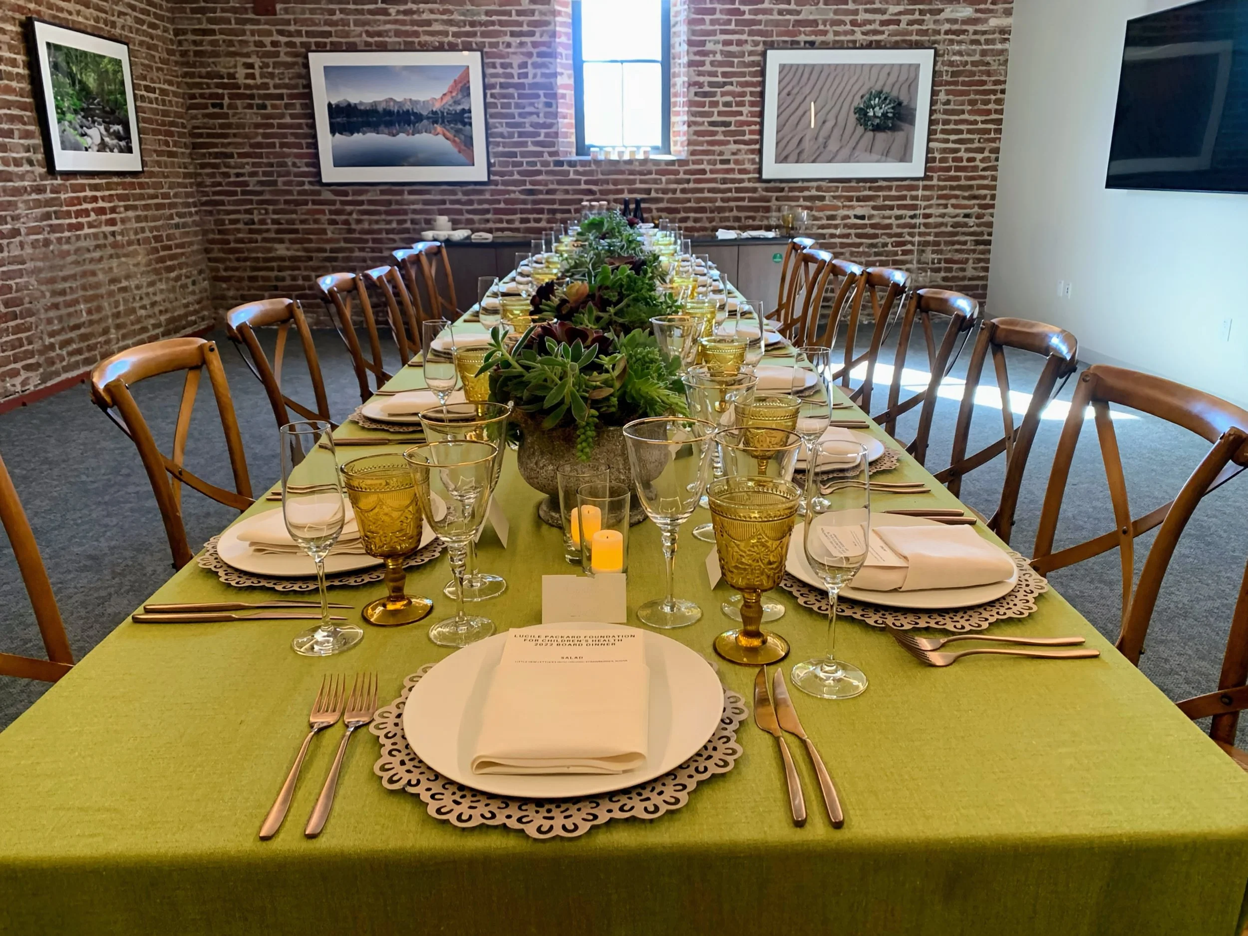 Long rectangular dining table with a green tablecloth set with white plates, napkins, silverware, and various glasses, with a centerpiece of succulents and small candles, in a room with brick walls and framed landscape photographs