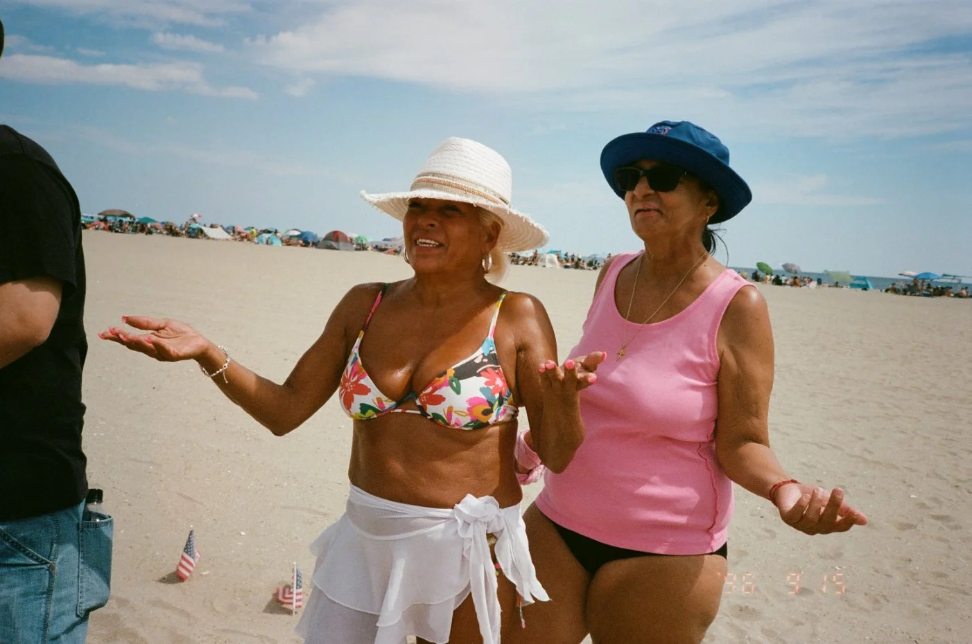 Two women standing on a sandy beach with many umbrellas and beachgoers in the background. One woman is wearing a floral bikini top, white skirt, and a wide-brimmed hat. The other woman is wearing a pink sleeveless top, black bottom, and a blue hat with sunglasses. Both women appear to be talking or gesturing.